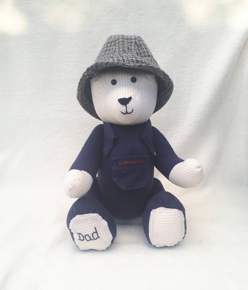 May include: A blue and white teddy bear wearing a tweed hat and a blue shirt with a pocket. The bear's foot is embroidered with the word "Dad".