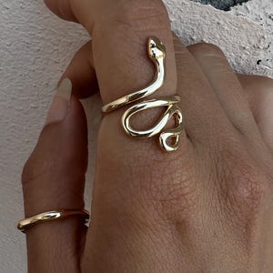 Könnte beinhalten: Ein goldfarbener Schlangenring umschlingt einen Finger, wobei sich der Schlangenkopf oben befindet. Ein einfacher goldener Bandring ist ebenfalls sichtbar. Die Ringe werden auf einer Hand vor einem strukturierten, cremefarbenen Hintergrund präsentiert.