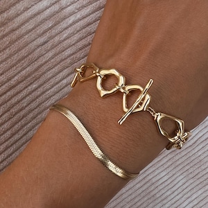 Pode incluir: Duas pulseiras de ouro no pulso de uma pessoa. A pulseira superior tem uma corrente grossa com elos em forma de coração. A pulseira inferior é uma corrente fina de cobra.