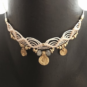 May include: A white and gold macrame choker necklace with a decorative pendant. The pendant is a circular design with a raised center and a textured surface. The necklace is accented with small gold beads and two gray stones.