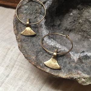 Könnte beinhalten: Ein Paar goldene Creolen mit kleinen axtförmigen Anhängern. Die Ohrringe liegen auf einer rauen, grauen Steinoberfläche.