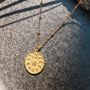 Puede incluir: Un collar de oro con un colgante redondo que presenta un diseño de sol y luna. El colgante está detallado con una estrella y lunas crecientes.