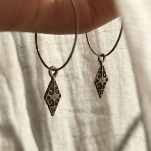Peut inclure: Une paire de boucles d'oreilles créoles en argent avec un délicat charme en forme de diamant orné d'un motif floral complexe.