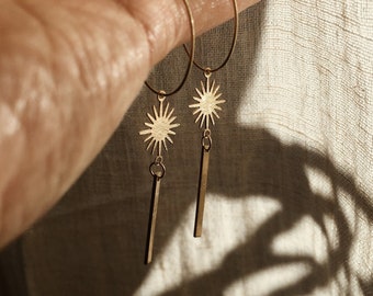 May include: A pair of gold hoop earrings with a starburst charm and a long, thin, dangling bar. The earrings are shown against a beige fabric background.