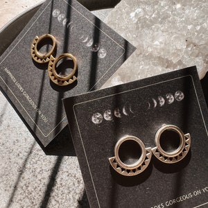 May include: Two pairs of gold hoop earrings with a geometric design. The earrings are displayed on black cards with the text "Happiness looks gorgeous on you".