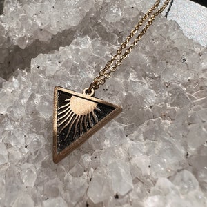 May include: A gold chain necklace with a triangular pendant featuring a sun design. The pendant is black and gold and has a textured surface.