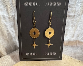 May include: Gold compass earrings with a star charm hanging below each compass. The earrings are displayed on a black card with a moon phase design.