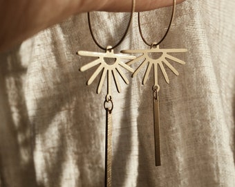 May include: Gold-toned hoop earrings with a sunburst design and a dangling rectangular bar. The earrings are held up against a neutral-colored fabric background. The earrings are a stylish accessory.