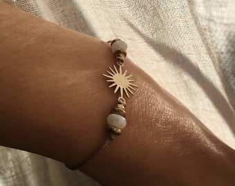 May include: A bracelet featuring a central gold-toned sunburst charm flanked by two white, semi-translucent beads and small gold-toned spacers. The charm is attached to a thin, neutral-colored cord that wraps around the wrist.