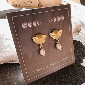 Puede incluir: Pendientes de oro con un diseño de sol y una perla colgante. Los pendientes se muestran en una tarjeta marrón con el texto "Happiness looks gorgeous on you."