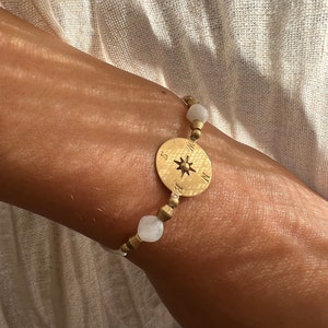 May include: A gold bracelet with white beads and a compass charm. The compass charm has a star in the center and the letters N, S, E, and W around the edge.