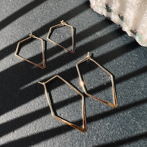 Puede incluir: Cuatro pendientes de aro geométricos de oro con forma de diamante. Los pendientes están sobre una superficie negra con luz y sombra.