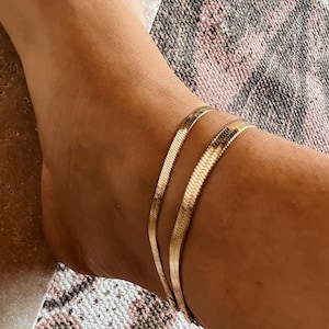 Könnte beinhalten: Zwei goldene Fußkettchen an einem Knöchel. Die Fußkettchen bestehen aus einer dünnen, flachen Kette mit leicht strukturierter Oberfläche.