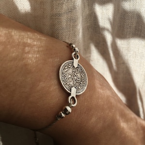 Puede incluir: Un brazalete de plata con un dije redondo en forma de moneda con escritura &aacute;rabe. El brazalete est&aacute; hecho de cord&oacute;n fino y peque&ntilde;as cuentas de plata.