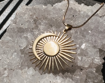 May include: A gold necklace with a sun and moon pendant. The pendant is a stylized sun with rays and a crescent moon shape. The necklace is on a bed of white crystals.