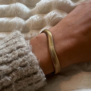 May include: A gold-colored, herringbone-style bracelet. The bracelet is worn on a wrist, with a gray sweater sleeve visible. The background is a soft, white, textured surface.