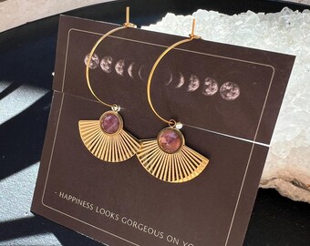 May include: Gold hoop earrings with fan-shaped pendants and a central pink gemstone. The earrings are displayed on a dark brown card with the text "- HAPPINESS LOOKS GORGEOUS ON YOU -" and a moon phase illustration.