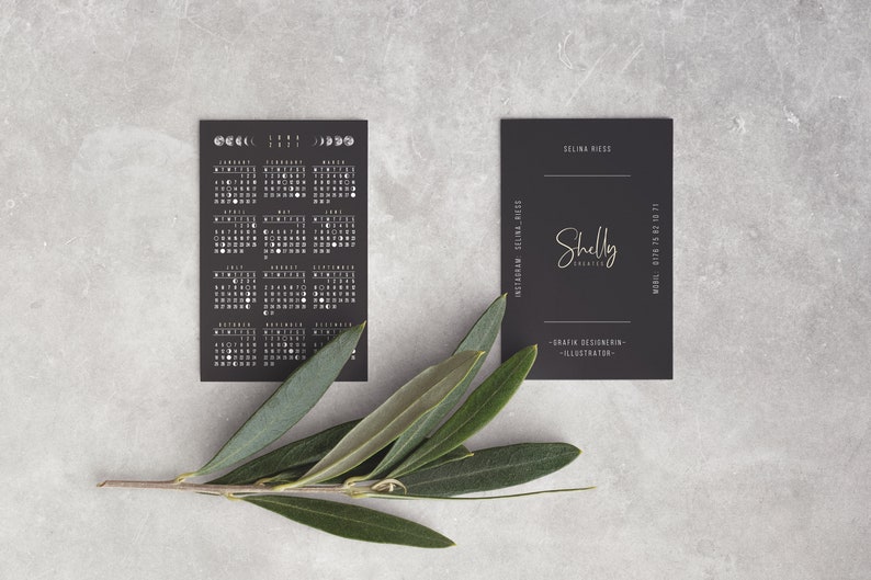 May include: Black and white calendar card with moon phases for each month of the year. A black business card with the text "Shelly Creates" and "Graphic Designer - Illustrator" is next to the calendar card.