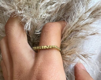 May include: A gold-toned ring with a textured, rectangular design. The ring is worn on a finger.