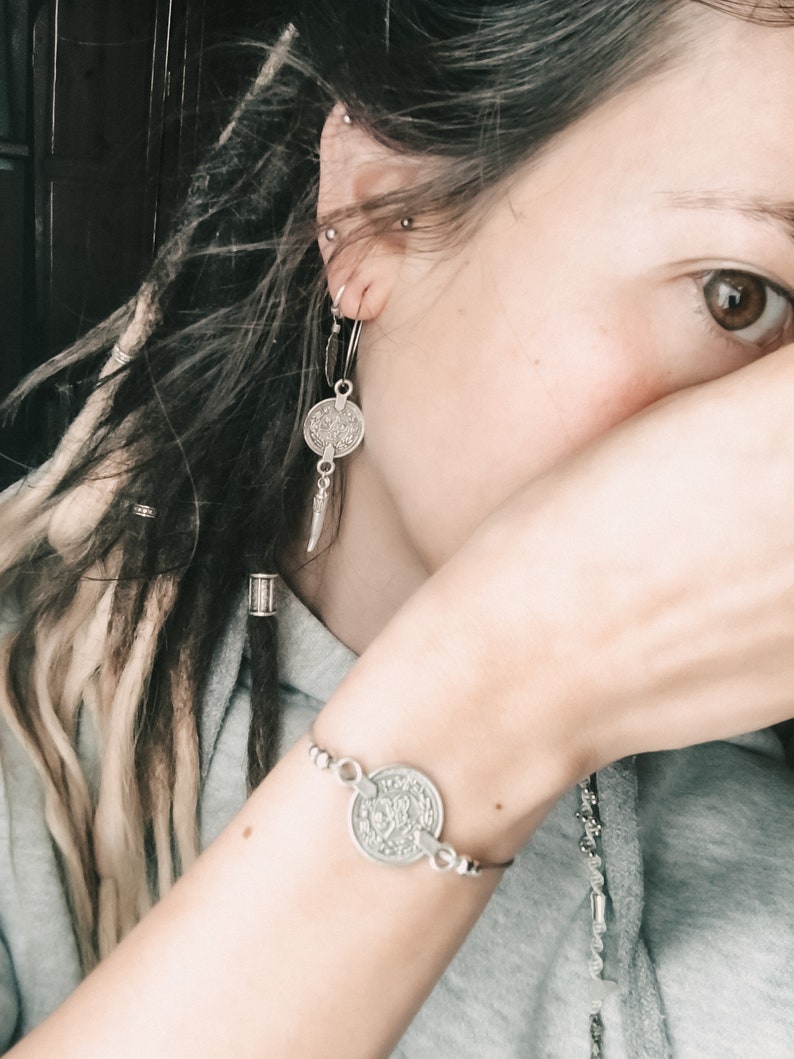 Puede incluir: Una pulsera de plata con un dije de moneda con dise&ntilde;os intrincados. La pulsera se lleva en la mu&ntilde;eca.