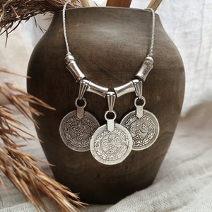 Puede incluir: Un collar de plata con tres colgantes redondos con diseños intrincados. El collar está hecho de un cordón blanco y cuentas de plata. El collar se muestra en un jarrón de cerámica marrón.