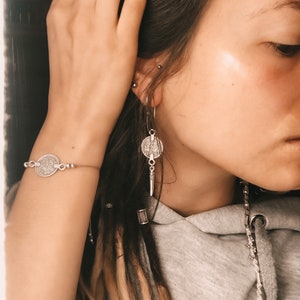 Puede incluir: Una persona con rastas largas marrones y rubias lleva un pendiente de moneda de plata, una pulsera de moneda de plata y un collar de cadena de plata. La persona lleva una sudadera gris.
