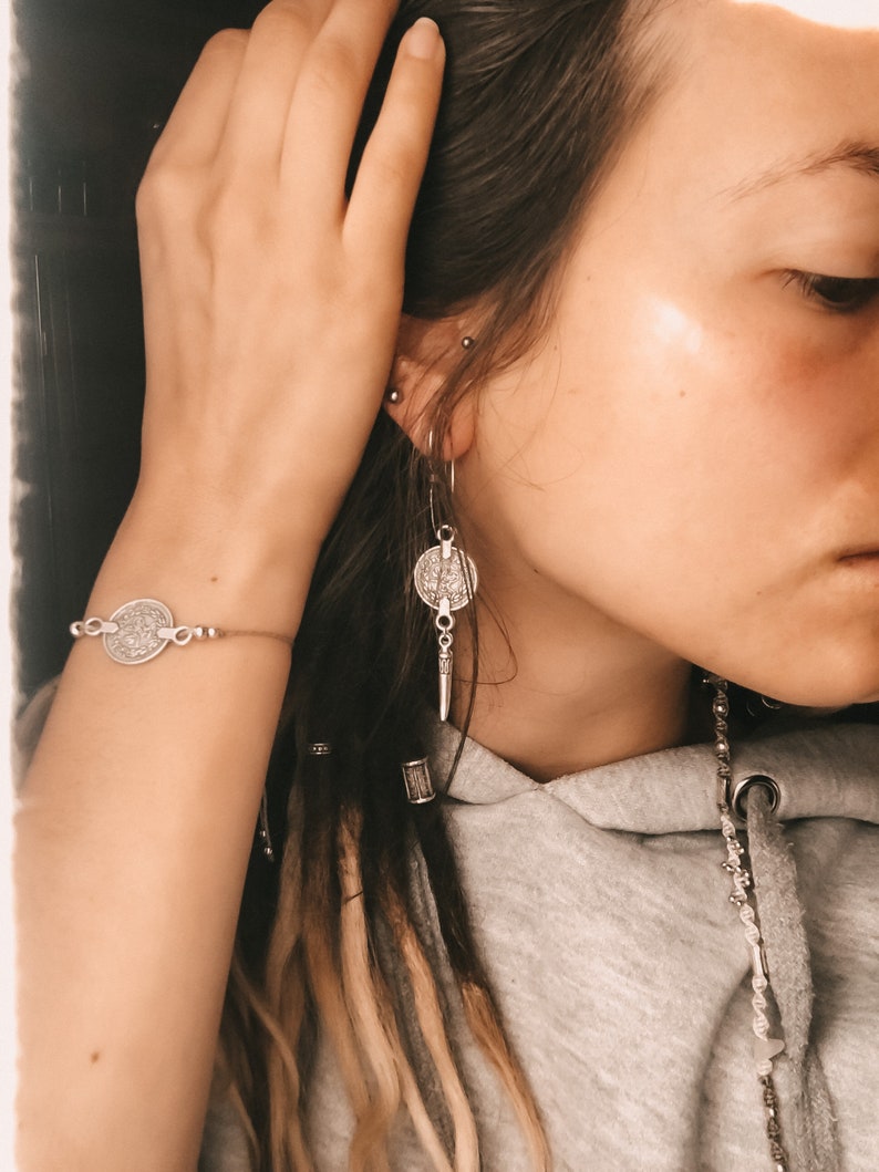 Puede incluir: Una persona con rastas largas marrones y rubias lleva un pendiente de moneda de plata, una pulsera de moneda de plata y un collar de cadena de plata. La persona lleva una sudadera gris.