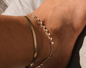 May include: Two gold anklets on a person's ankle. The top anklet is a delicate chain with small round charms. The bottom anklet is a thicker, flat chain.