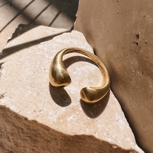 Peut inclure: Une bague en métal doré avec une forme unique en forme de larme. La bague est ouverte et a une finition lisse et polie.