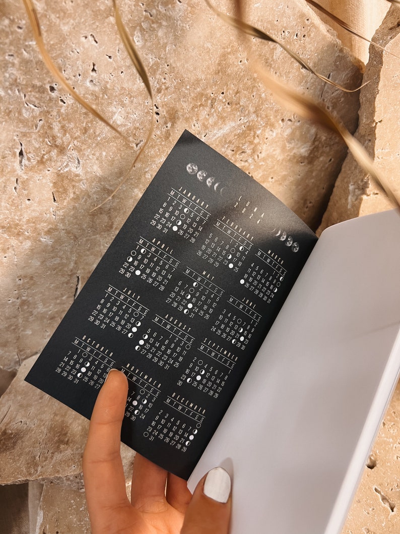 Puede incluir: Un calendario en blanco y negro con las fases lunares para cada d&iacute;a del a&ntilde;o 2024. El calendario est&aacute; abierto a los meses de julio y agosto.