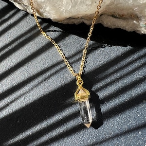 May include: A gold-colored necklace with a clear crystal pendant. The crystal is shaped like a pointed prism. The necklace chain is delicate and hangs against a dark, textured surface.