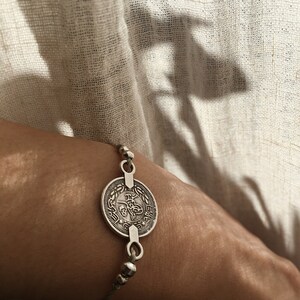 Puede incluir: Un brazalete de plata con un gran dije de moneda con escritura &aacute;rabe. El brazalete est&aacute; en la mu&ntilde;eca de una persona.