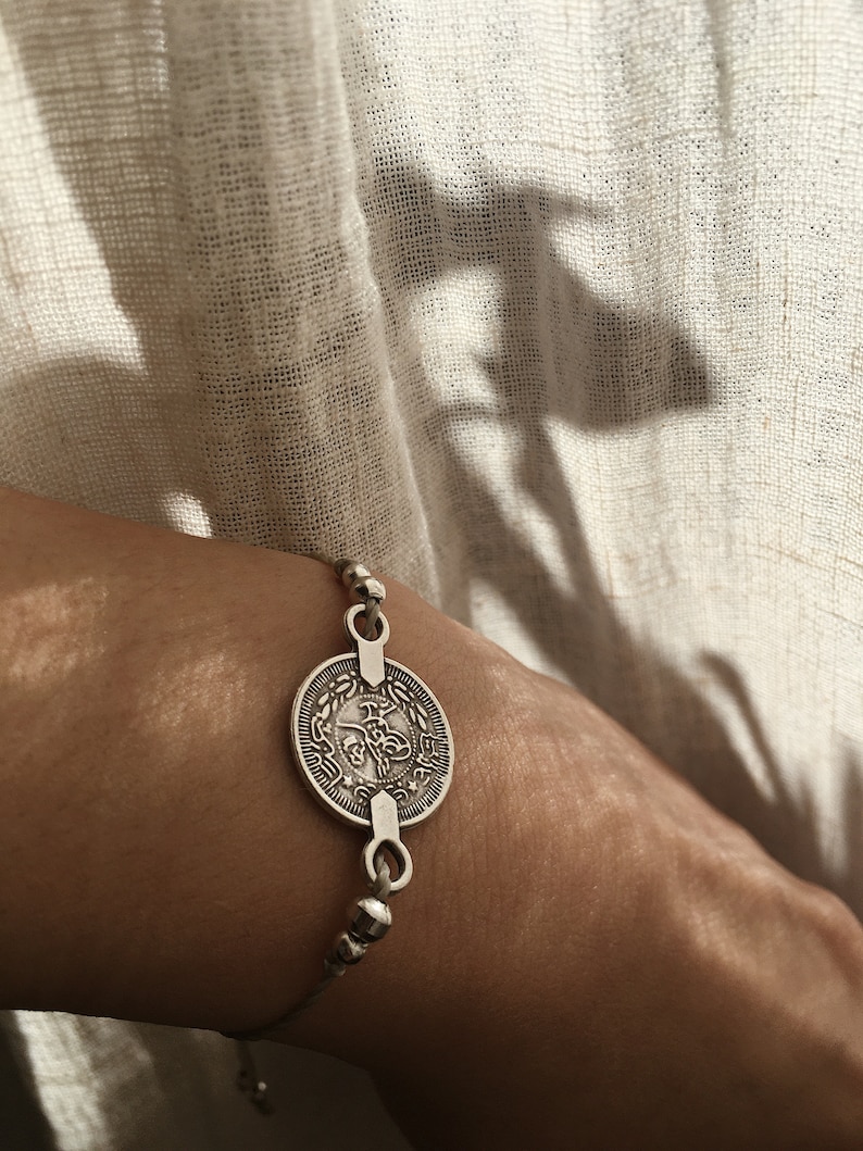 Puede incluir: Un brazalete de plata con un gran dije de moneda con escritura &aacute;rabe. El brazalete est&aacute; en la mu&ntilde;eca de una persona.