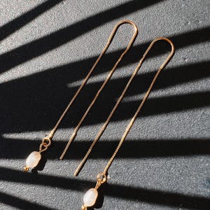 Op de afbeelding: Een paar gouden kettingoorbellen met een kleine parelbedel aan elke oorbel.