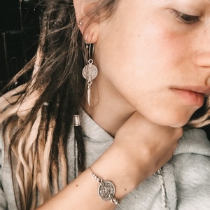 Puede incluir: Una pulsera de plata con un dije de moneda. La pulsera est&aacute; en la mu&ntilde;eca de una persona. La persona tiene rastas largas y rubias y lleva una sudadera gris.