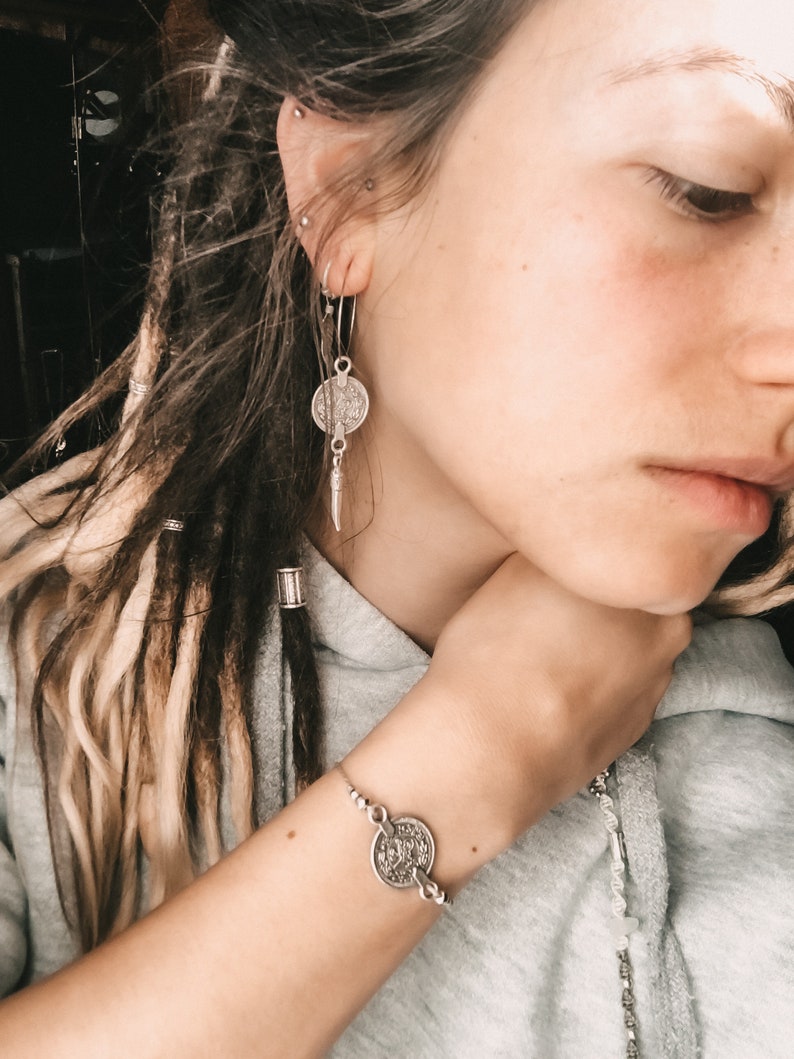 Puede incluir: Una pulsera de plata con un dije de moneda. La pulsera est&aacute; en la mu&ntilde;eca de una persona. La persona tiene rastas largas y rubias y lleva una sudadera gris.