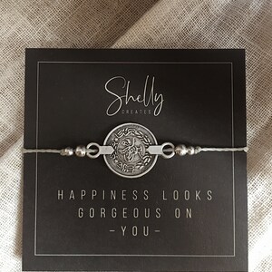 Puede incluir: Un brazalete de plata con un dije de moneda que dice "La felicidad se ve hermosa en ti" en una tarjeta negra con el texto "Shelly Creates" en ella.