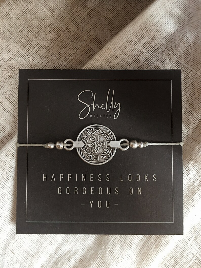 Puede incluir: Un brazalete de plata con un dije de moneda que dice "La felicidad se ve hermosa en ti" en una tarjeta negra con el texto "Shelly Creates" en ella.