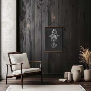 May include: A black and white anatomical illustration of a human skeleton in a seated pose, printed on a poster hanging on a dark wood wall. The poster is framed with a black frame and hung with a black hanging system. The wall is in a room with a wooden floor and a white chair.