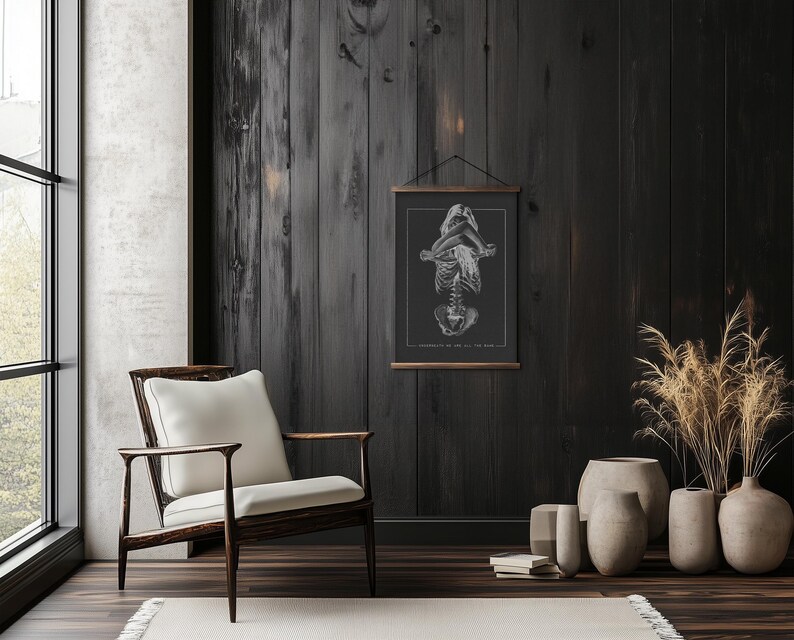 May include: A black and white anatomical illustration of a human skeleton in a seated pose, printed on a poster hanging on a dark wood wall. The poster is framed with a black frame and hung with a black hanging system. The wall is in a room with a wooden floor and a white chair.