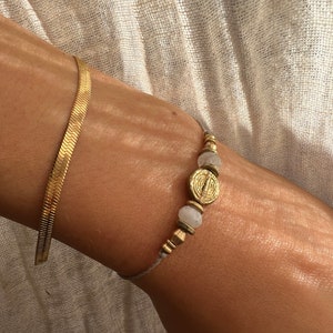 Op de afbeelding: Een gouden kettingarmband met een grijs koord en witte en gouden kralen. De armband heeft een gouden bedel met een cirkelvormig ontwerp.