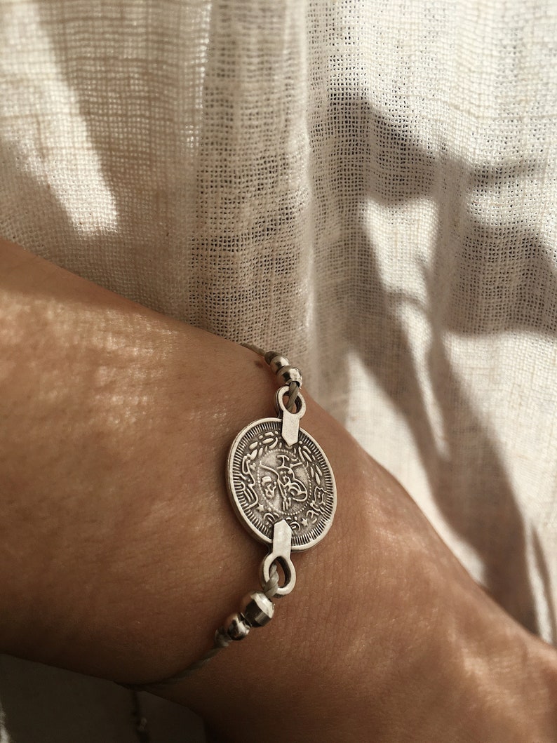 Puede incluir: Un brazalete de plata con un dije redondo en forma de moneda con escritura &aacute;rabe. El brazalete est&aacute; hecho de cord&oacute;n fino y peque&ntilde;as cuentas de plata.