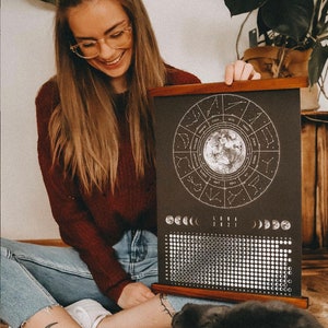 May include: A black and white moon phase calendar with a wooden frame. The calendar features a circle with the zodiac signs and a moon in the center. The calendar also includes a grid of moon phases for the year 2021.