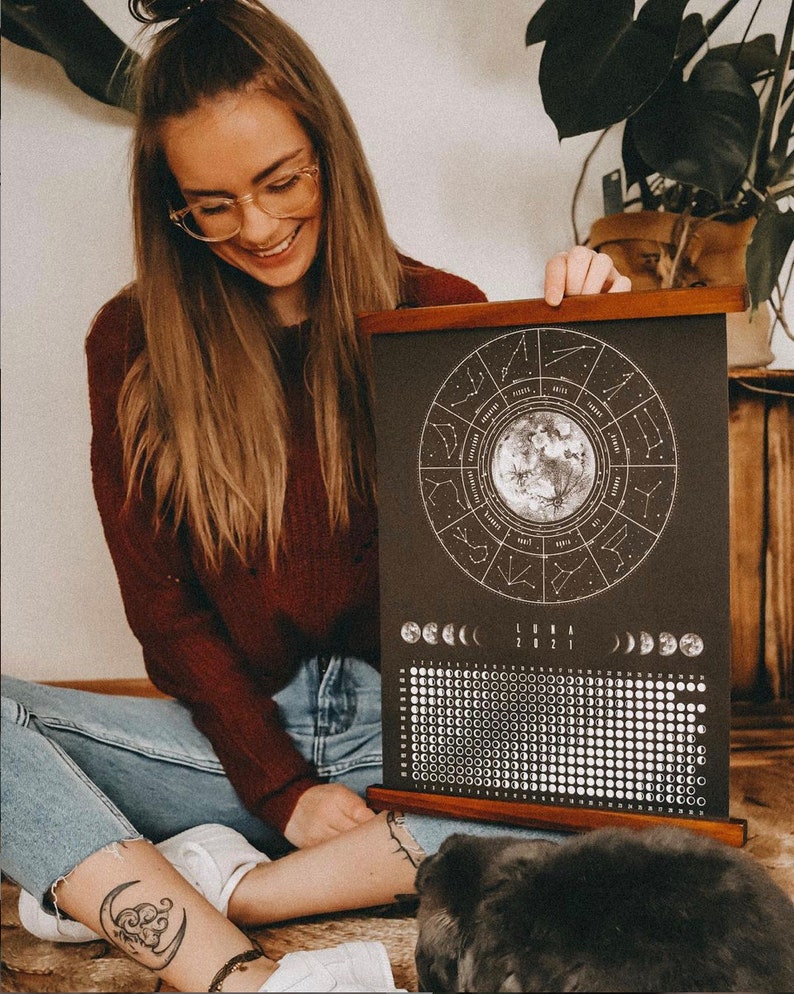May include: A black and white moon phase calendar with a wooden frame. The calendar features a circle with the zodiac signs and a moon in the center. The calendar also includes a grid of moon phases for the year 2021.