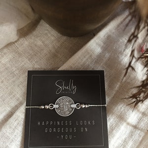 Puede incluir: Un brazalete plateado con un dije redondo que dice "La felicidad se ve hermosa en ti." El brazalete est&aacute; sobre una tarjeta negra con las palabras "Shelly Creates" impresas en ella.