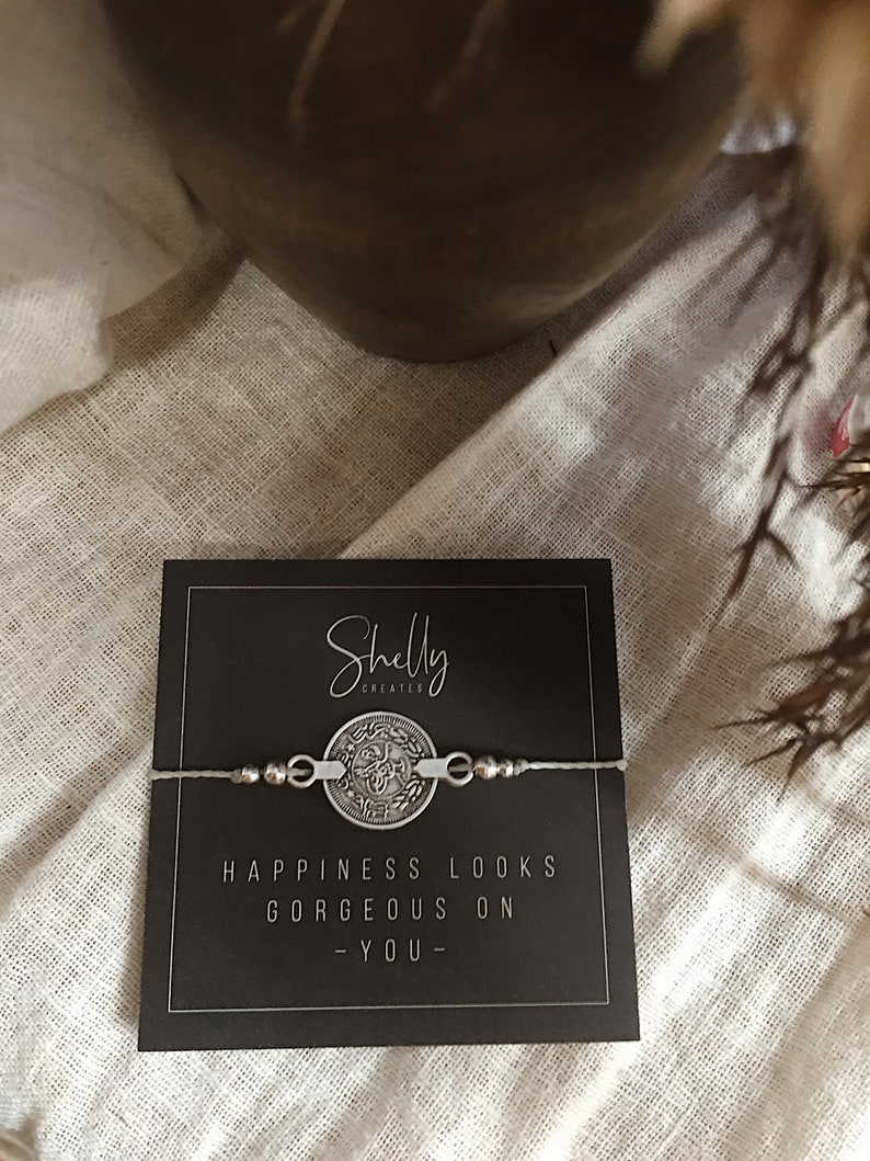 Puede incluir: Un brazalete plateado con un dije redondo que dice "La felicidad se ve hermosa en ti." El brazalete est&aacute; sobre una tarjeta negra con las palabras "Shelly Creates" impresas en ella.