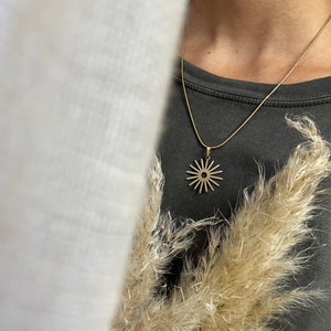 Op de afbeelding: Een gouden ketting met een zonvormige hanger. De hanger heeft een cirkelvormig midden en stralende lijnen.