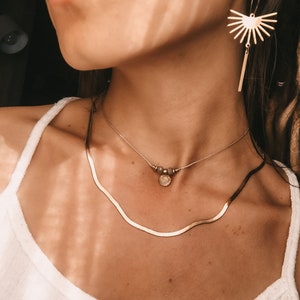 以下が含まれることがあります： 白いタンクトップを着た女性が、金のチェーンネックレスと金のペンダントをつけています。彼女はまた、太陽の光が放射状に広がったデザインの金のイヤリングもつけています。