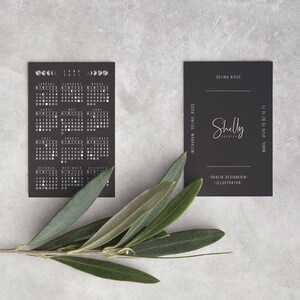Puede incluir: Tarjeta de calendario en blanco y negro con fases lunares y una tarjeta de visita negra con el texto "Shelly Creates" y "Dise&ntilde;adora gr&aacute;fica - Ilustradora" en letras doradas.