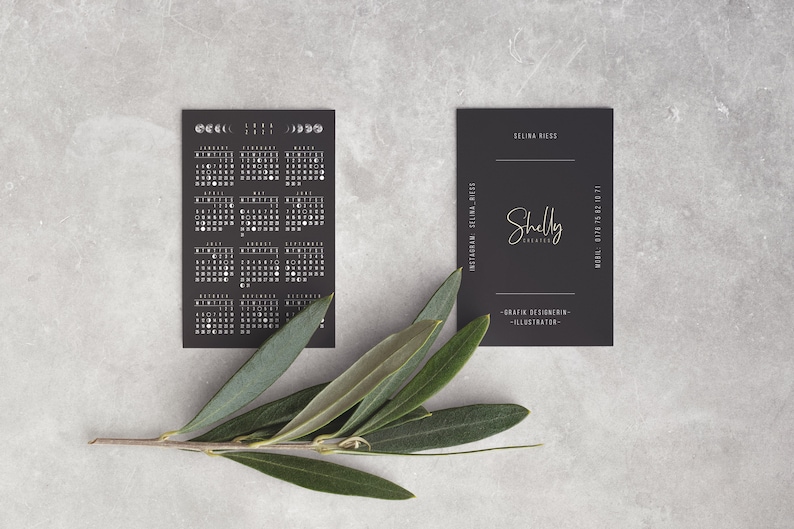 Puede incluir: Tarjeta de calendario en blanco y negro con fases lunares y una tarjeta de visita negra con el texto "Shelly Creates" y "Dise&ntilde;adora gr&aacute;fica - Ilustradora" en letras doradas.